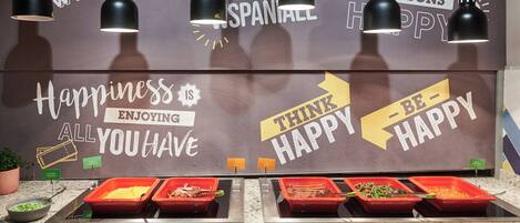 Zona de bufé con múltiples bandejas de comida y una pared decorativa con frases como “La felicidad es todo lo que tienes” y “Piensa en positivo”.