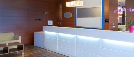 Una zona de recepción con pared de madera, mostrador blanco, lámpara colgante moderna y una orquídea en maceta.