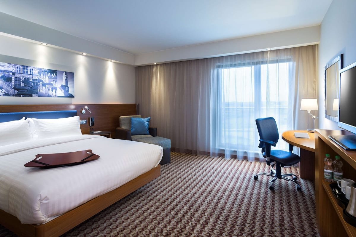 Une chambre d’hôtel avec un grand lit, un bureau avec une télévision, une chaise, une petite table et une fenêtre avec des rideaux.