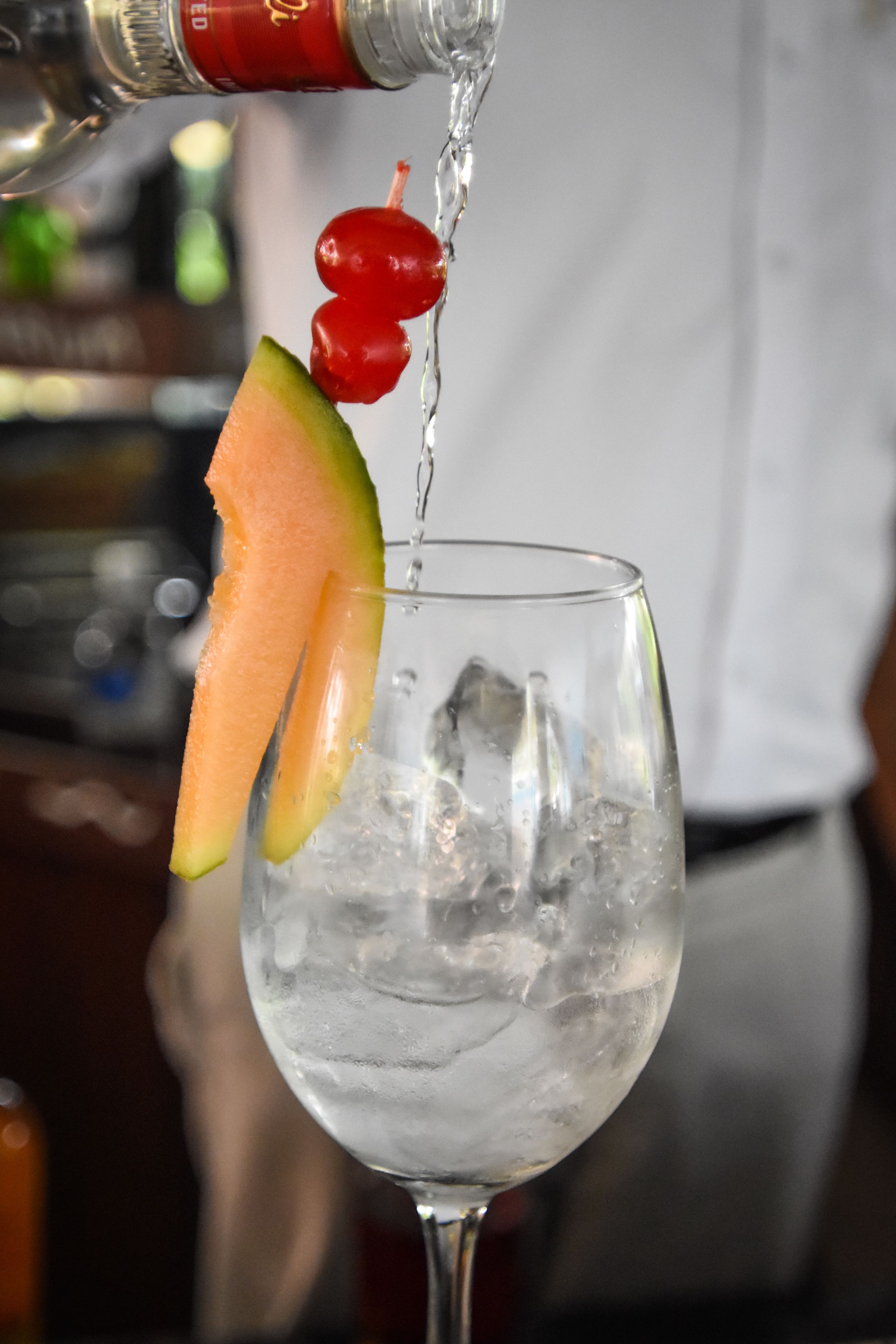 Een cocktail wordt over ijs in een glas geschonken, versierd met een schijfje meloen en een kers.