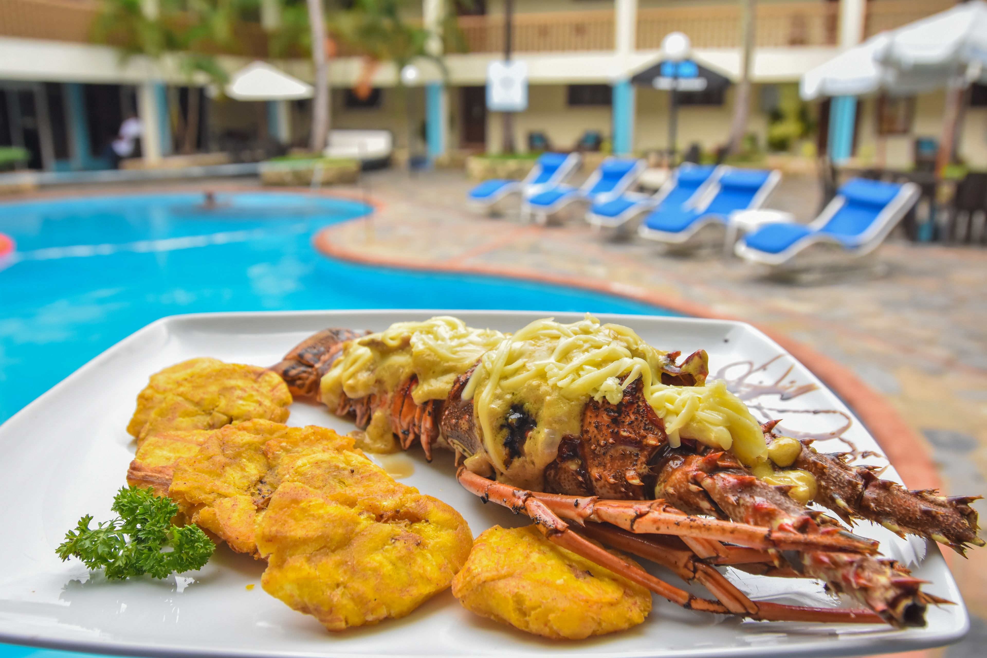 A lobster dish with yellow sauce and fried plantains on a plate, set against a poolside background with lounge chairs and umbrellas.