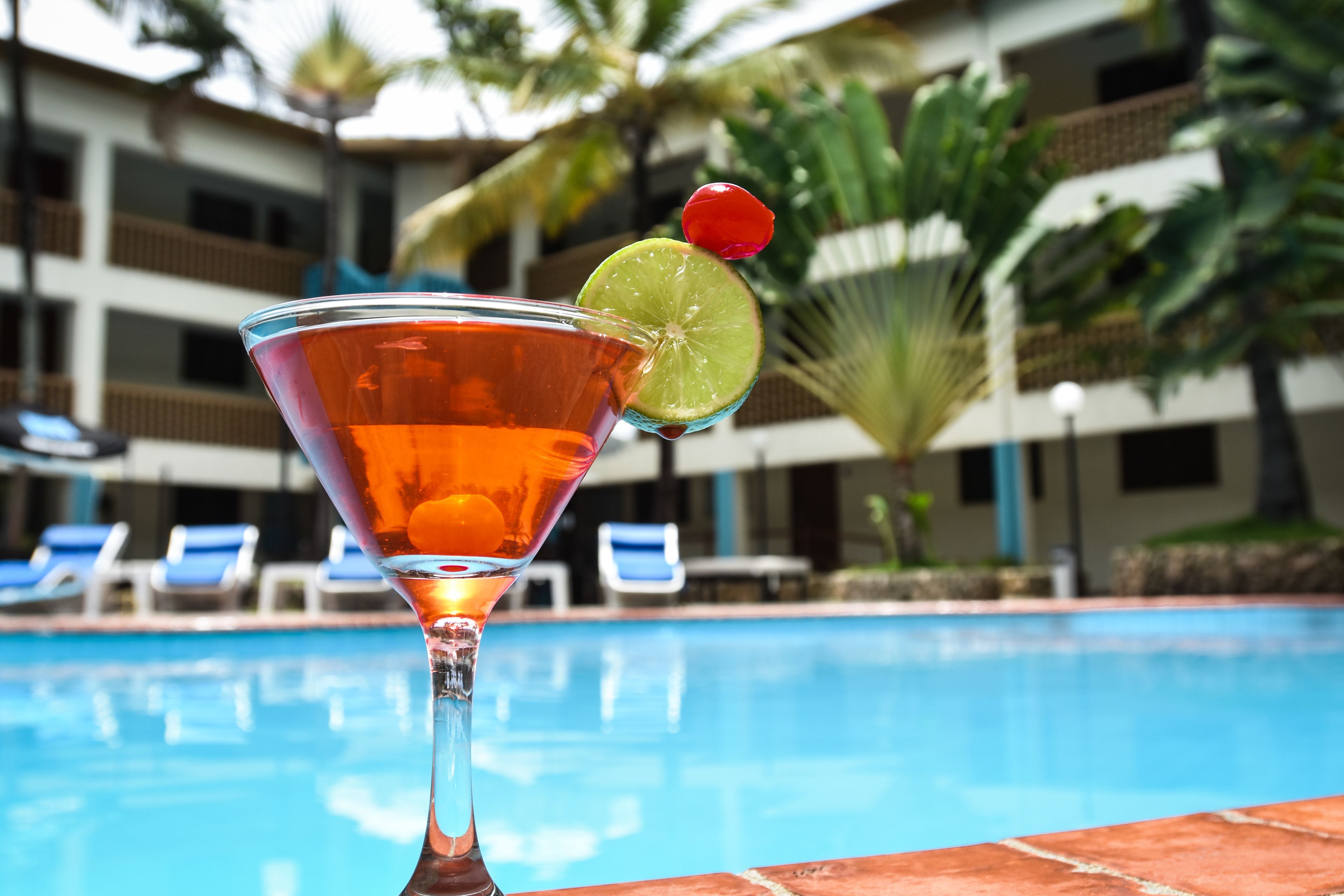 En cocktail med et limeskive og en kirsebærpynt i et martiniglas ved en pool, med en hotelbygning i baggrunden.