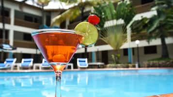 Un cocktail, agrémenté d’une tranche de citron vert et d’une cerise, servi dans un verre à martini au bord d’une piscine, avec un bâtiment d’hôtel en arrière-plan.