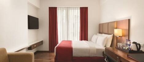 Une chambre d’hôtel avec un lit, un bureau, une chaise, une télévision et une fenêtre avec des rideaux.