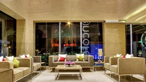 Un hall d’entrée moderne avec un ensemble de canapés de couleur beige, une table basse et une grande fenêtre qui reflète les lumières et un bâtiment.