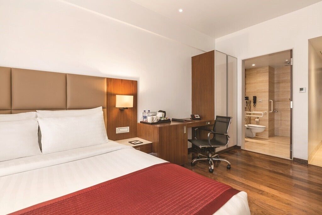 Habitación de hotel con cama, escritorio, silla y baño visible a través de una puerta abierta.