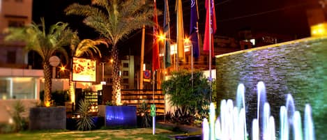 A nighttime view of a hotel courtyard with palm trees, a fountain, and flags.