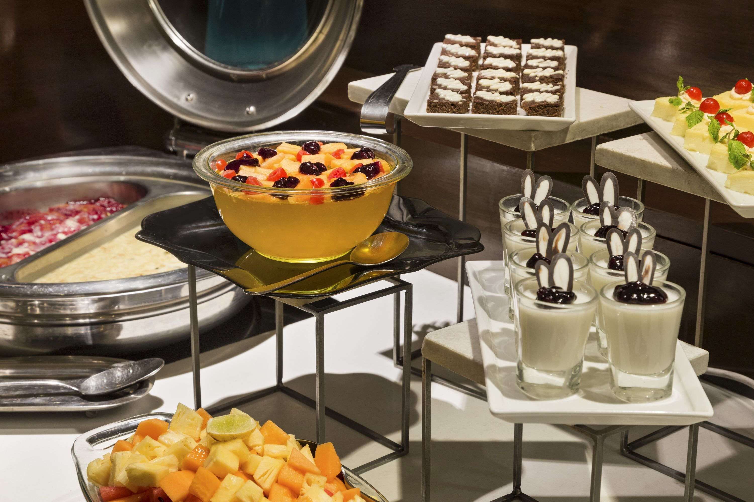 Un bufé con diversos alimentos, incluyendo un tazón de ensalada de frutas, una bandeja de postres y varios recipientes de vidrio con dulces.