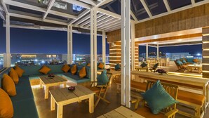 Un espace de détente à aire ouverte, comprenant des sièges et des tables, de grandes fenêtres et une vue sur le paysage urbain illuminé la nuit.