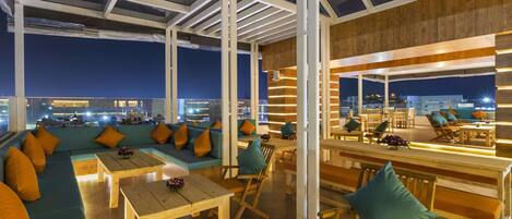 Un salón tipo loft con asientos y mesas, ventanas amplias y vistas a la ciudad iluminada de noche.