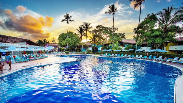 A swimming pool with lounge chairs and umbrellas, surrounded by palm trees and buildings. - Resort Arcobaleno All Inclusive (Porto Seguro)