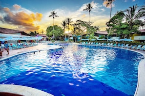 A swimming pool with lounge chairs and umbrellas, surrounded by palm trees and buildings. - Resort Arcobaleno All Inclusive (Porto Seguro)