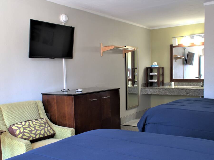 Habitación de hotel con dos camas, un televisor montado en la pared, un tocador de madera, una silla con cojín estampado, un espejo y una mesita.