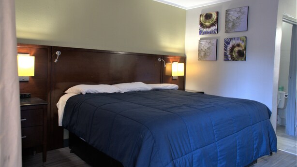 A neatly made bed with a dark blue bedspread and white pillows, a wooden headboard, two bedside lamps, and four wall-mounted artwork pieces. - Eastern Shore Motel (Daphne)