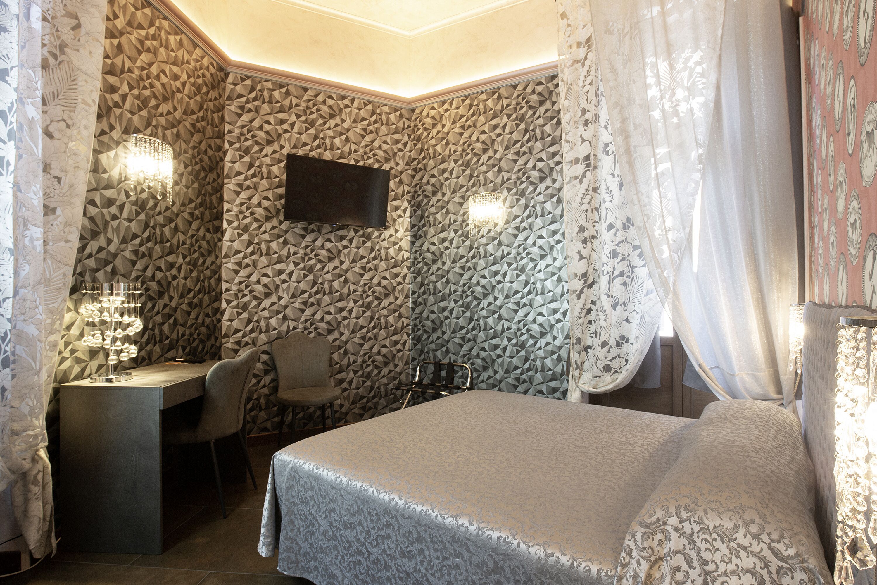A hotel room with a geometric-patterned wall, a flat-screen TV, a desk with a chair, and a bed with a patterned bedspread.