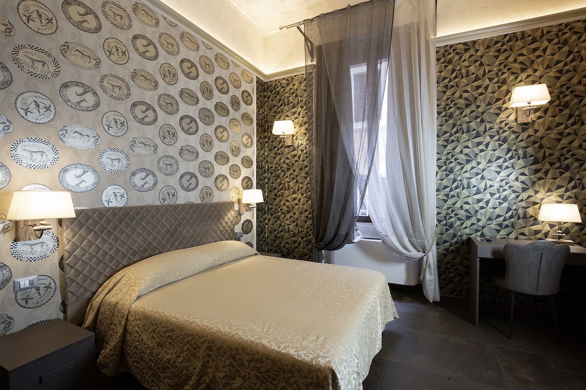 Habitación de hotel con cama, escritorio y una silla. Las paredes tienen papel tapiz estampado.