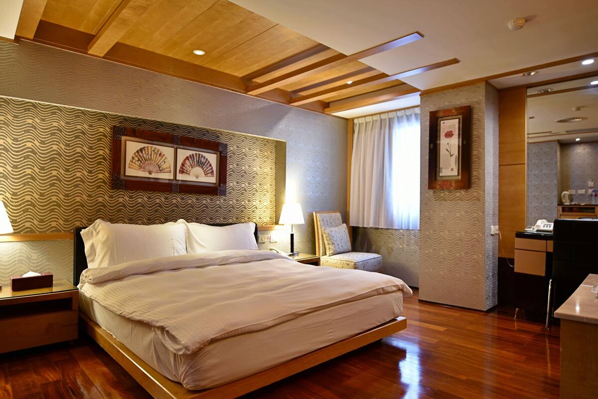A hotel room with a large bed, wooden flooring, a ceiling with exposed beams, and a small sitting area with a chair and table.