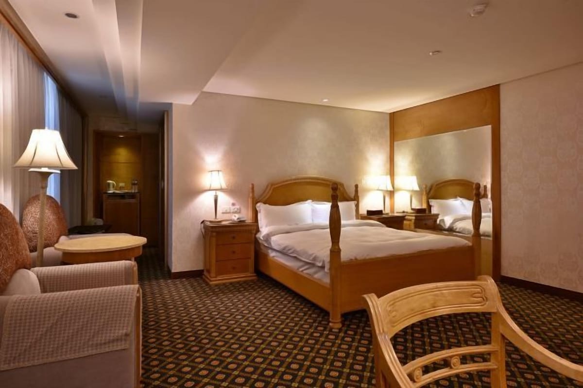 A hotel room with a four-poster bed, two bedside tables with lamps, a round table with a chair, and a large mirror on the wall.
