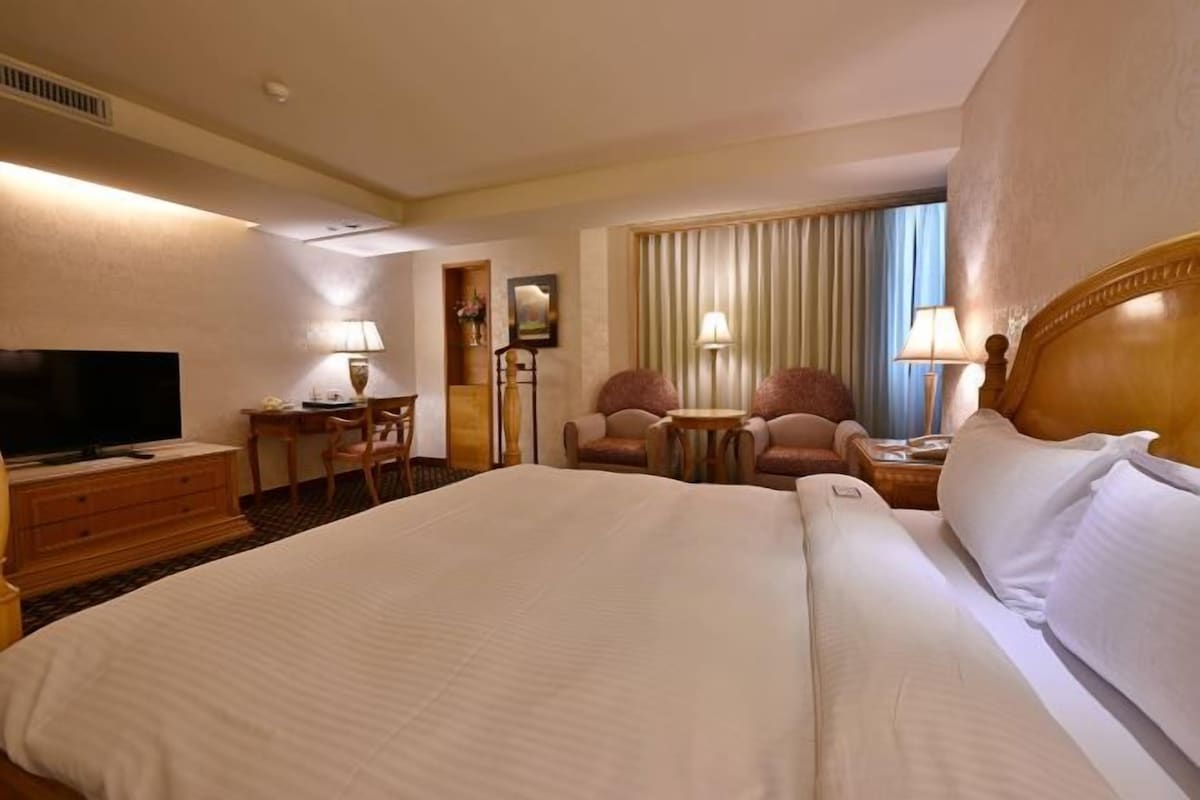 A hotel room with a large bed, a desk with a chair, a television, and a window with curtains.