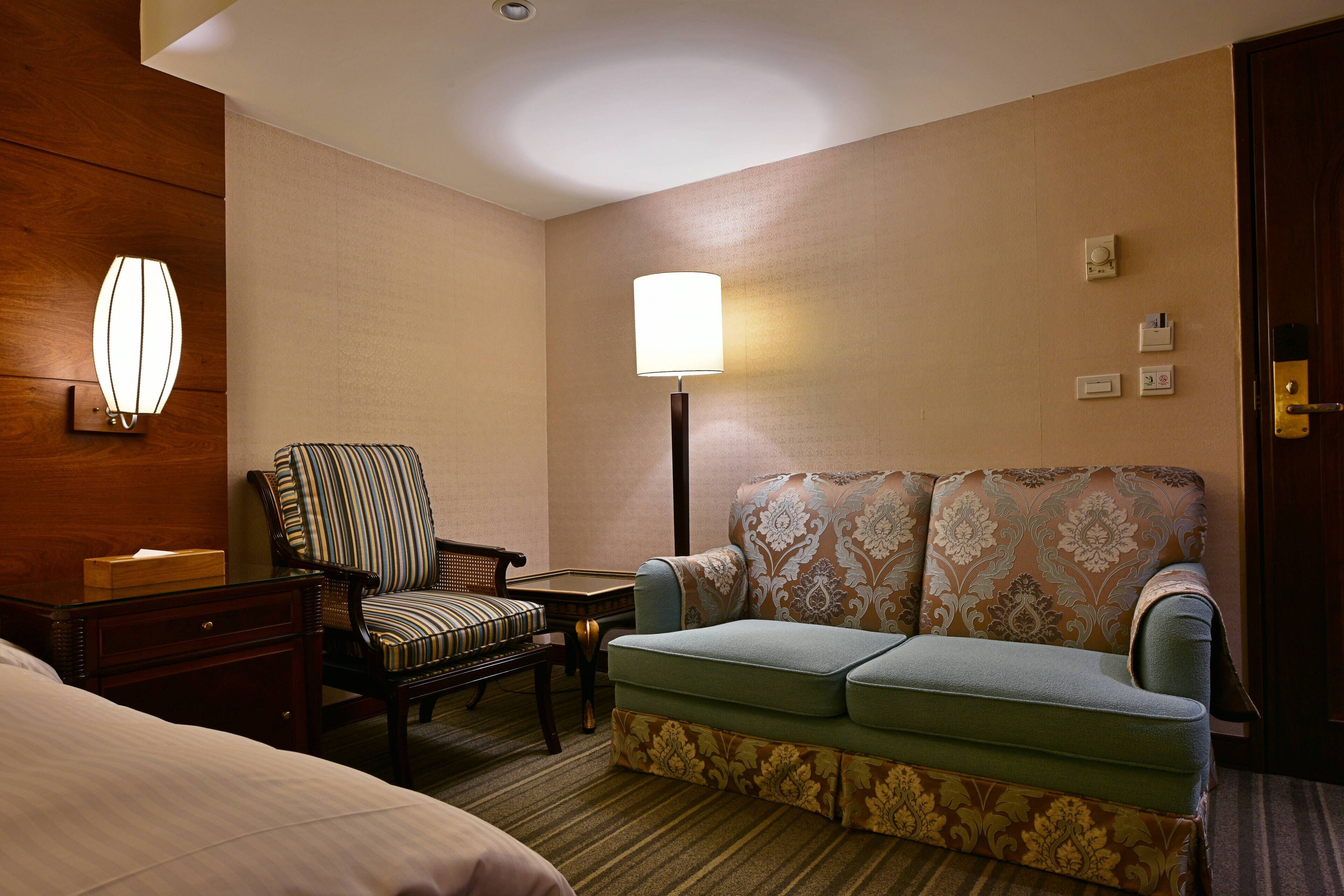A hotel room with a bed, a striped armchair, a patterned sofa, a small table, and a floor lamp.