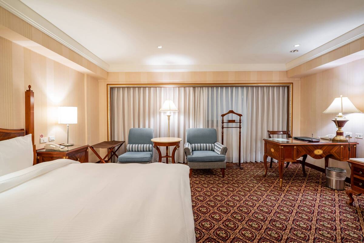 A hotel room with a large bed, two armchairs, a desk, a chair, a small table, and a patterned carpet.