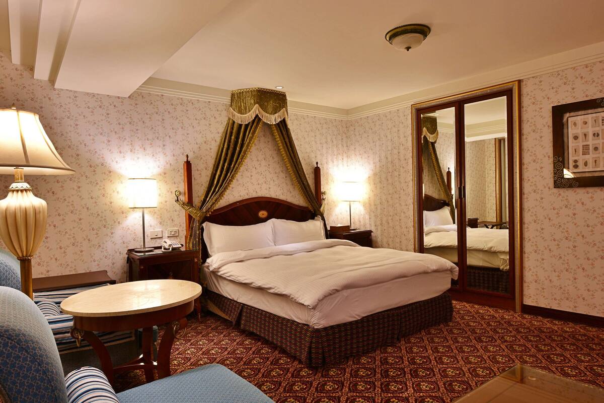 A hotel room with a large bed, two bedside tables with lamps, a chair, a small round table, a mirror, and a patterned carpet.