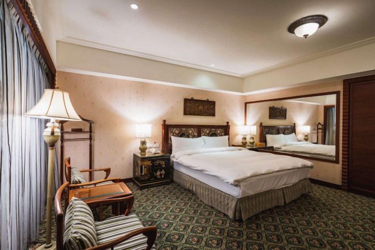 Une chambre d’hôtel avec un grand lit, deux tables de chevet avec des lampes, une chaise, une petite table et un grand miroir.