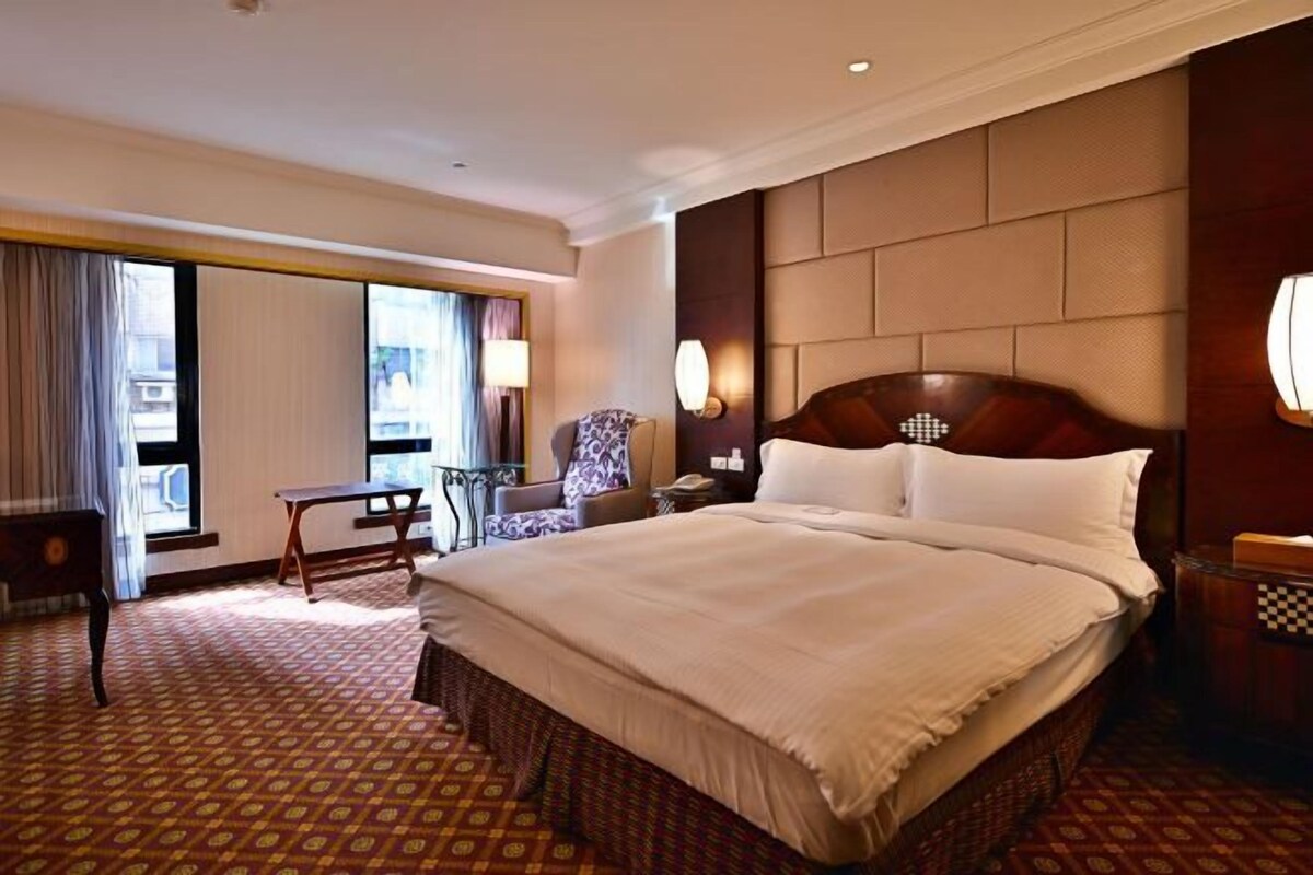 A hotel room with a large bed, a desk, a chair, and a view of the city through large windows.