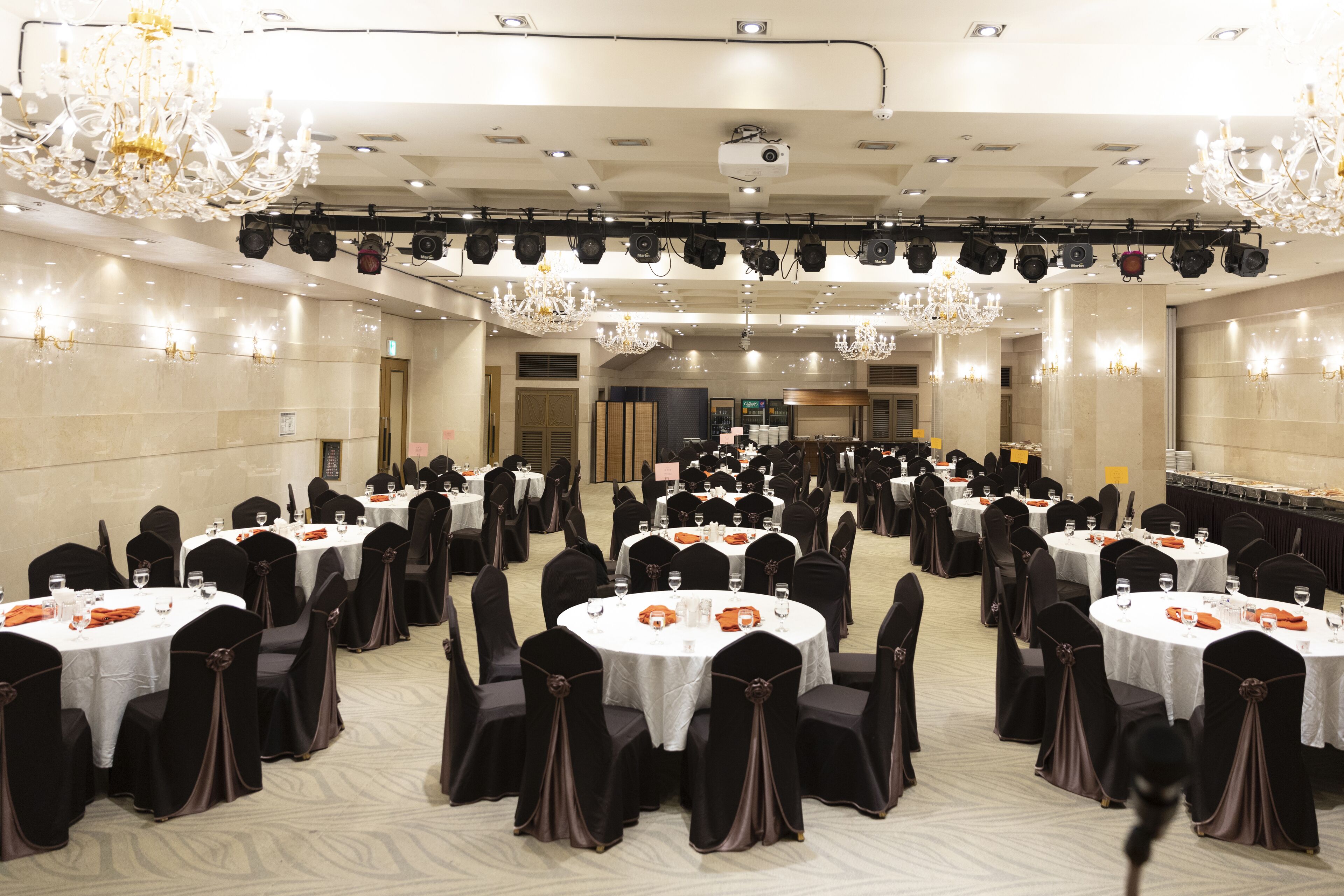A banquet hall with round tables, black chairs, and chandeliers.