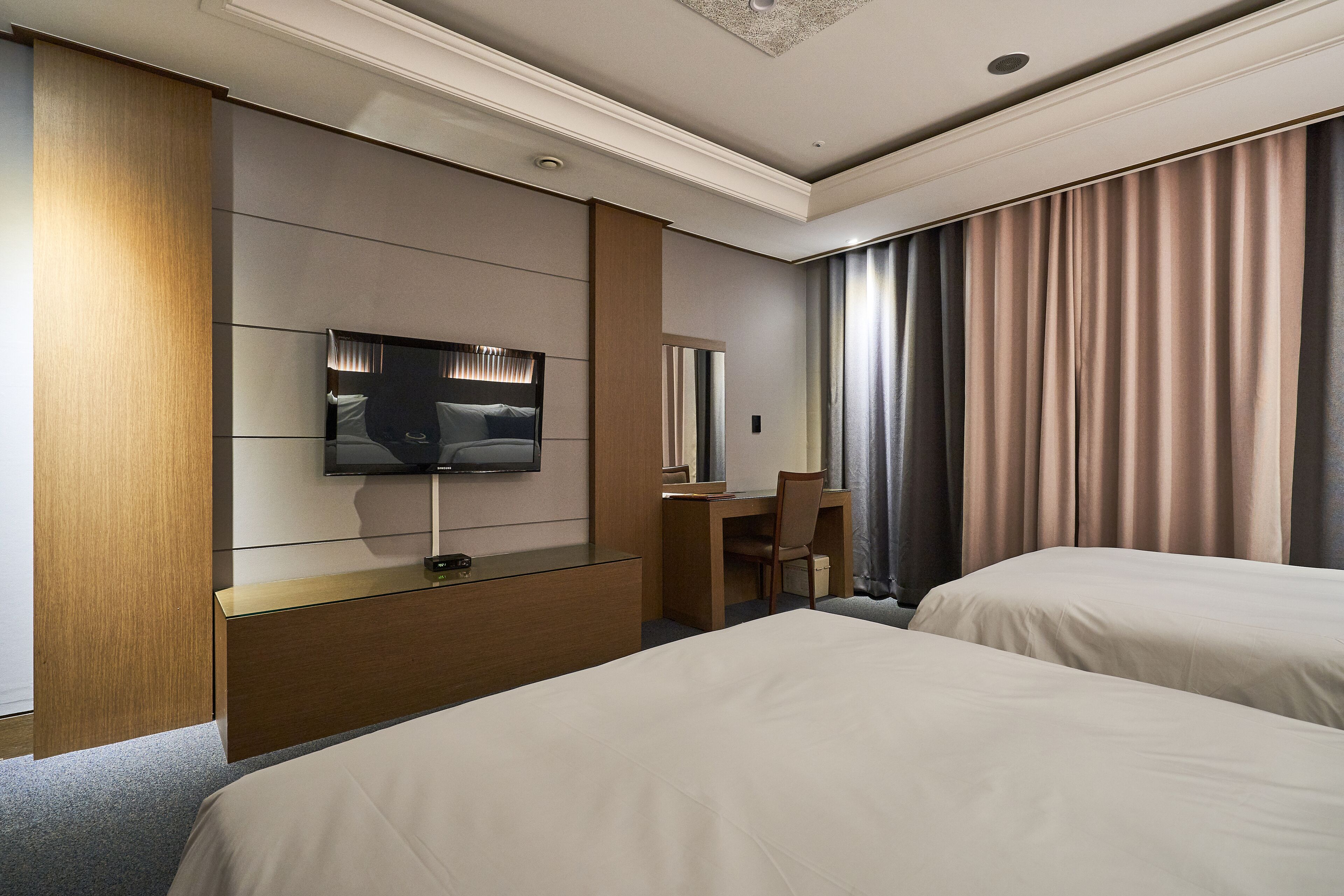 A hotel room with two beds, a desk, a chair, a TV, and a large window with curtains.