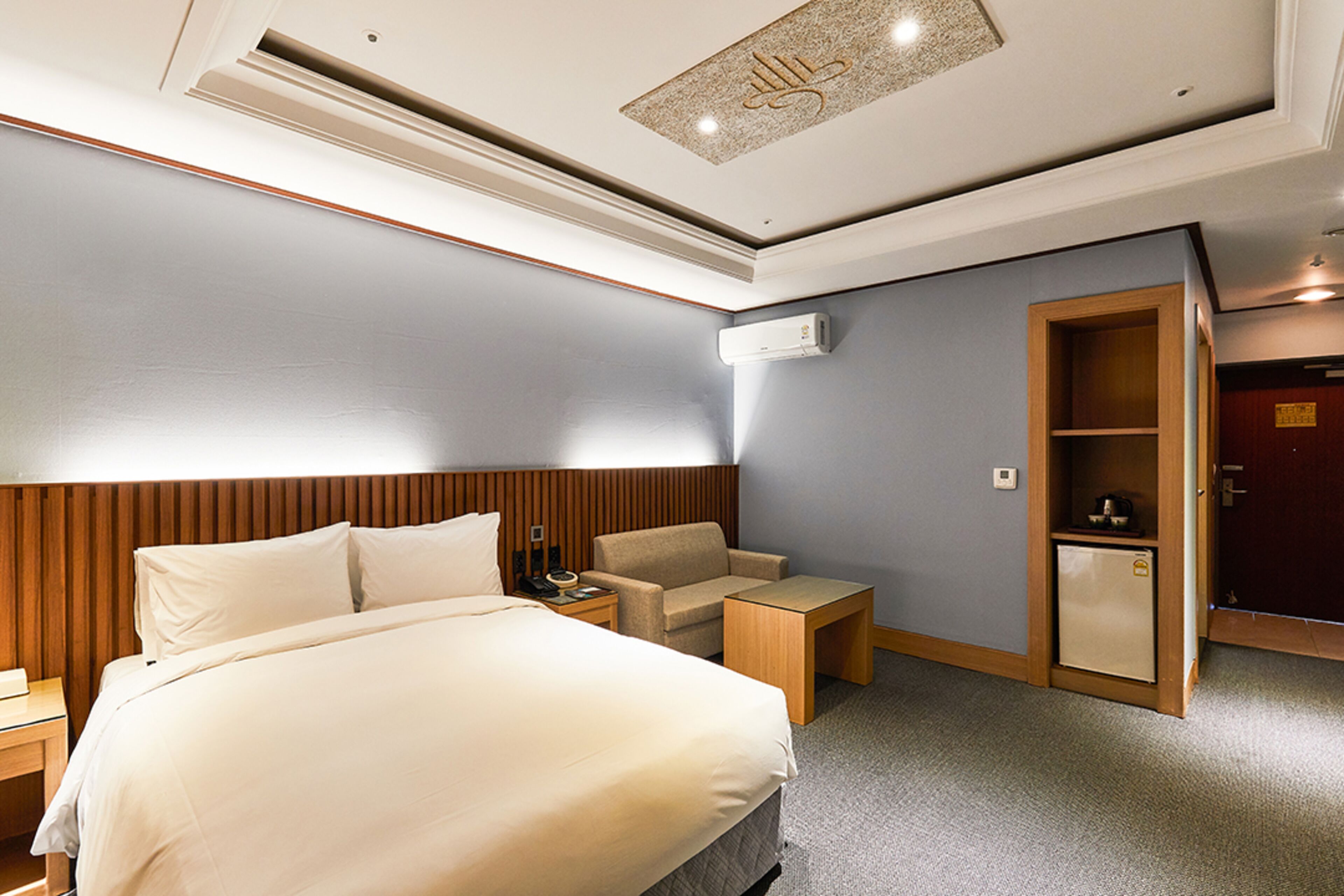A hotel room with a bed, a chair, a nightstand, a desk, a mini-fridge, and a wardrobe.