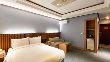 A hotel room with a bed, a chair, a nightstand, a desk, a mini-fridge, and a wardrobe.