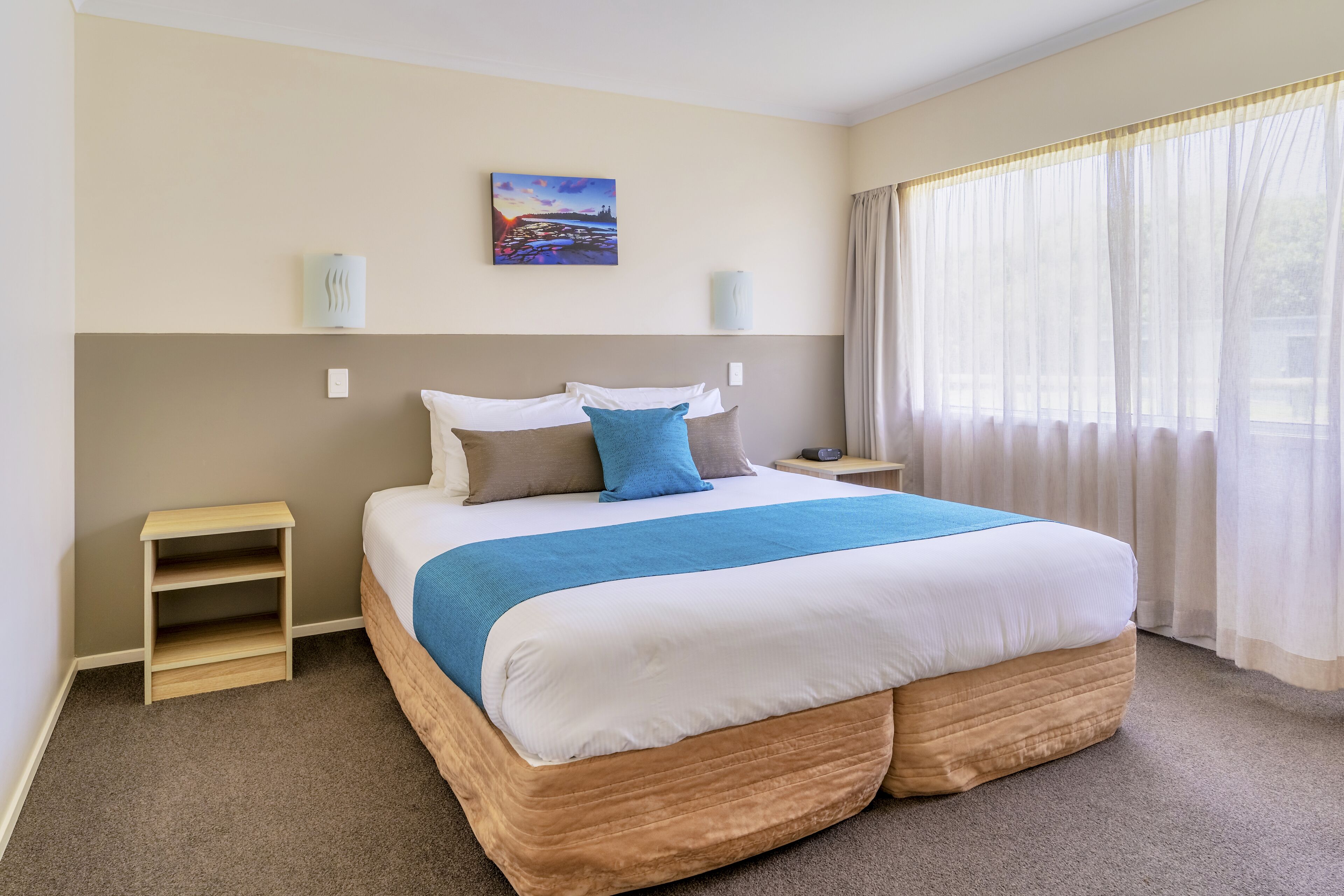 Habitación de hotel ordenada con una cama grande, una mesita de noche, un estante pequeño y una ventana con cortinas de tul.