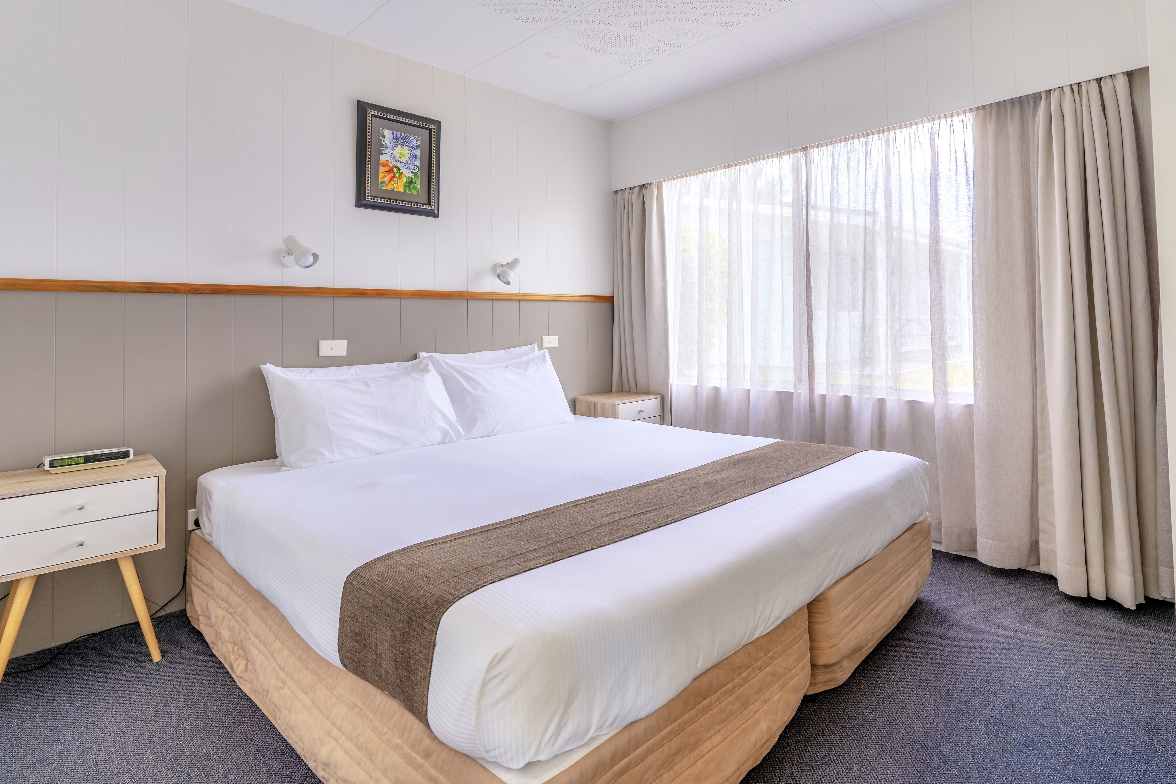 A hotel room with a large bed, a bedside table, a nightstand, a wall-mounted lamp, a window with curtains, and a framed picture on the wall.