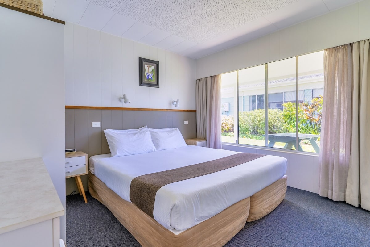 A hotel room with a large bed, a bedside table, a nightstand, a window with curtains, and a view of greenery outside.