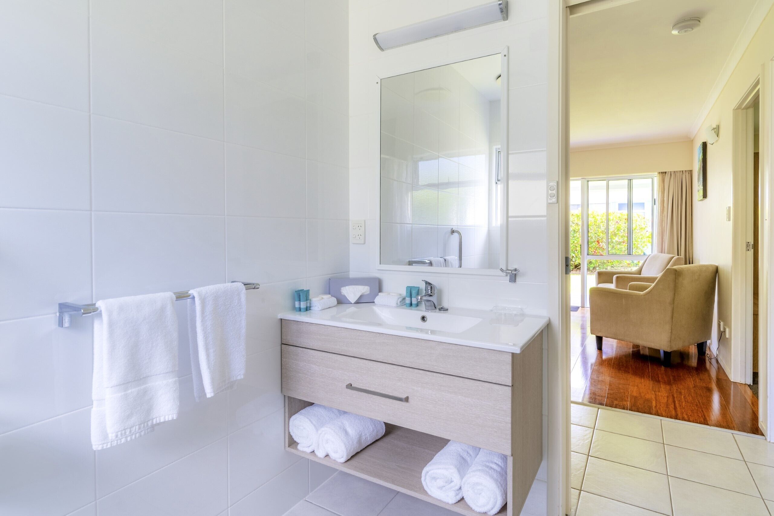 A modern bathroom with a white sink, mirror, and towel rack. There is a wooden vanity with drawers and a shelf for towels.