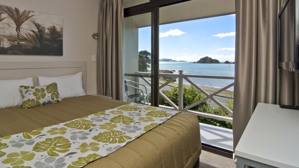 A hotel room with a bed, a painting on the wall, and a balcony with a view of the sea. - Breakwater Motel (Paihia)