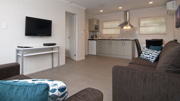 Una sala moderna con una televisión de pantalla plana, una mesa de centro y un sofá. Hay una zona de cocina con microondas, refrigerador y una ventana con persianas.