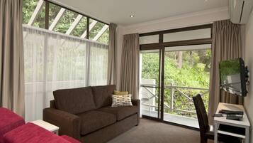 A room with a brown sofa, a small white table, a desk with a computer, and a chair. There is a large window with curtains and a view of greenery outside.