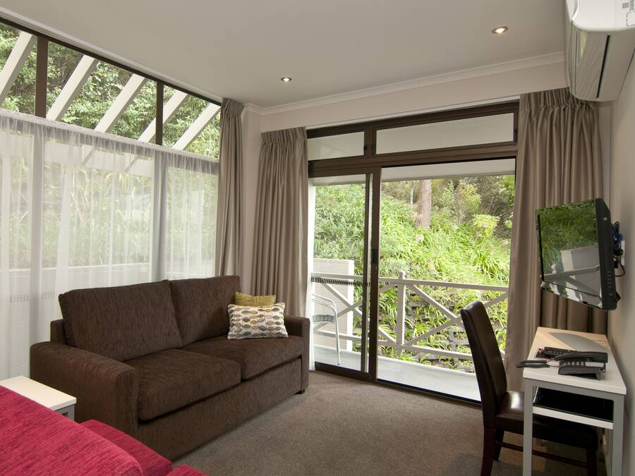 A room with a brown sofa, a small white table, a desk with a computer, and a chair. There is a large window with curtains and a view of greenery outside.