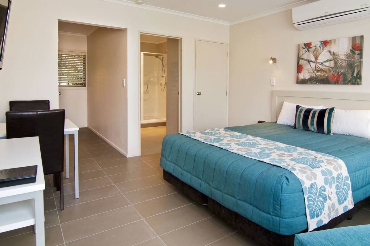 Quarto de hotel com cama, escrivaninha, cadeira, banheiro e uma pintura na parede.
