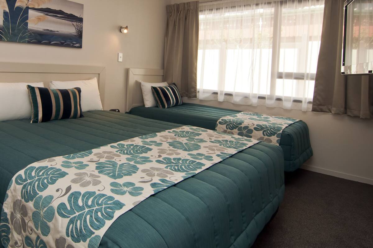 Quarto de hotel com duas camas, uma mesinha de cabeceira, um quadro na parede e uma janela com cortinas.