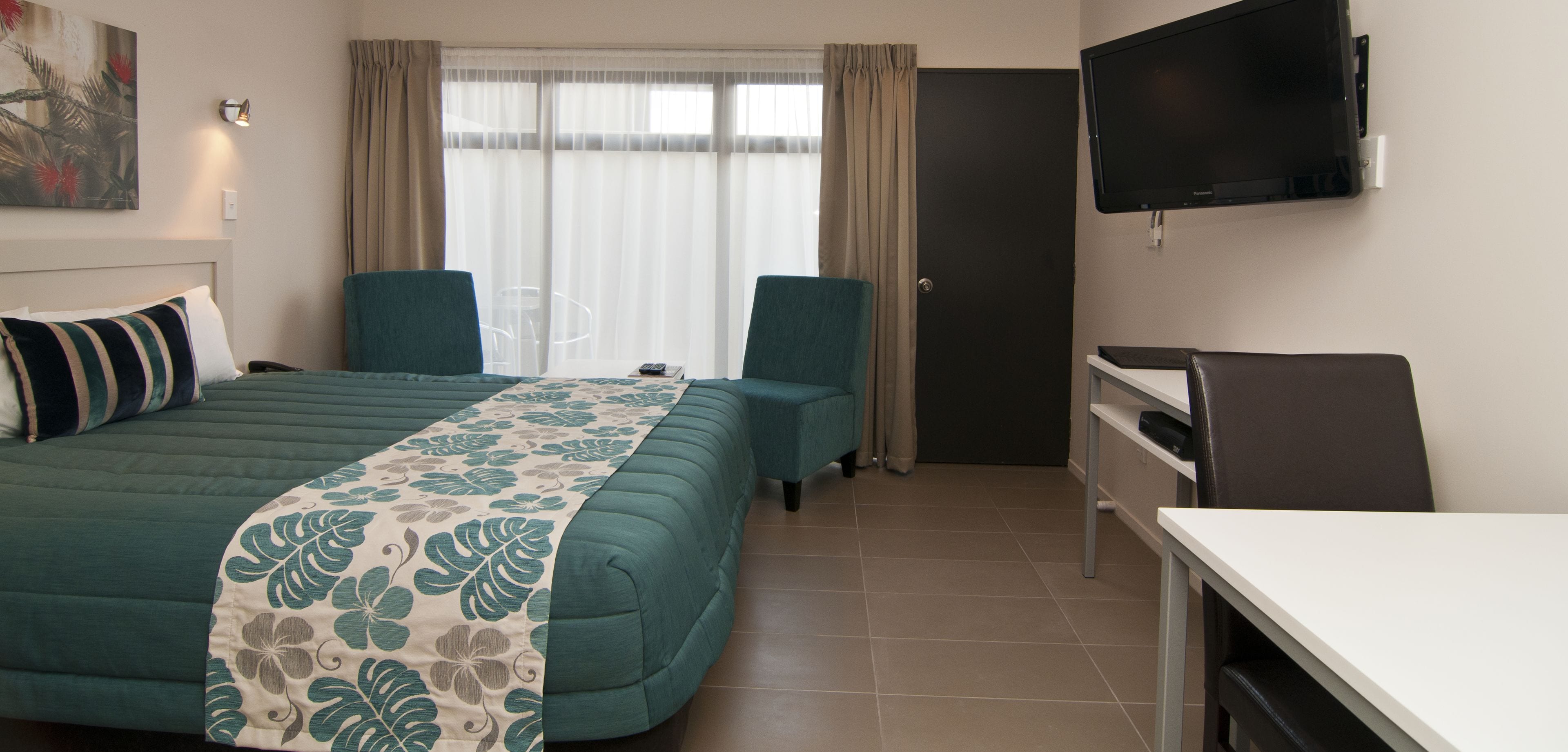A hotel room with a bed, two chairs, a desk, a TV, and a window with curtains.