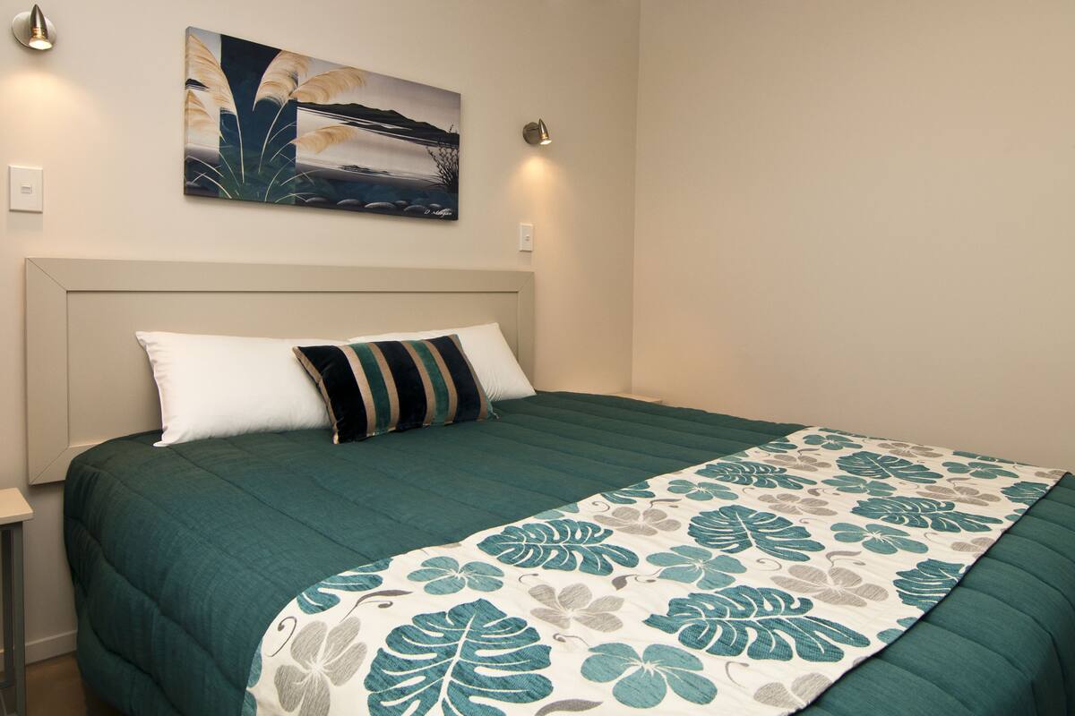 Cama bem arrumada com colcha floral verde e branca, um travesseiro branco e um travesseiro listrado. Há uma pintura sobre a cama e um luminária na parede à direita.