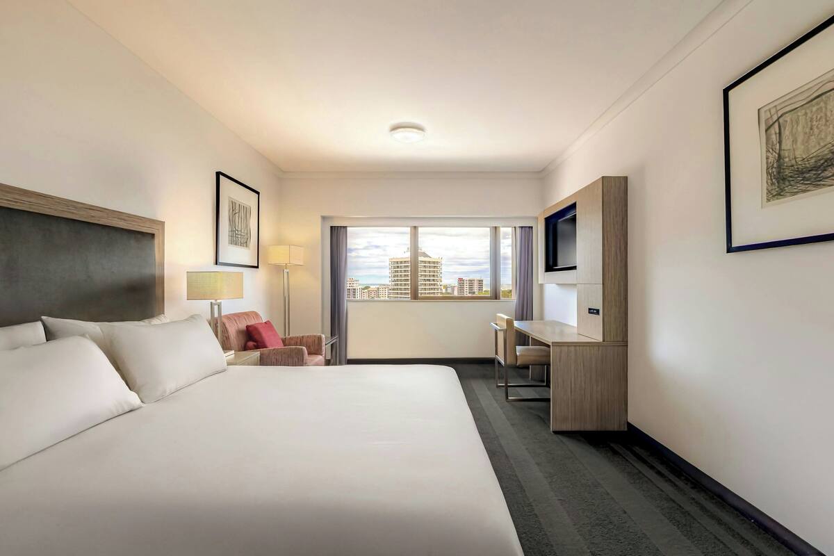 Habitación de hotel con una cama grande, un escritorio, una silla, un televisor y una ventana con vista a la ciudad.
