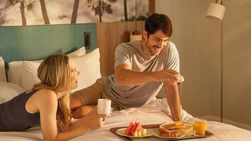A couple in a hotel room with a breakfast tray, bed, pillows, and a wall with palm trees.