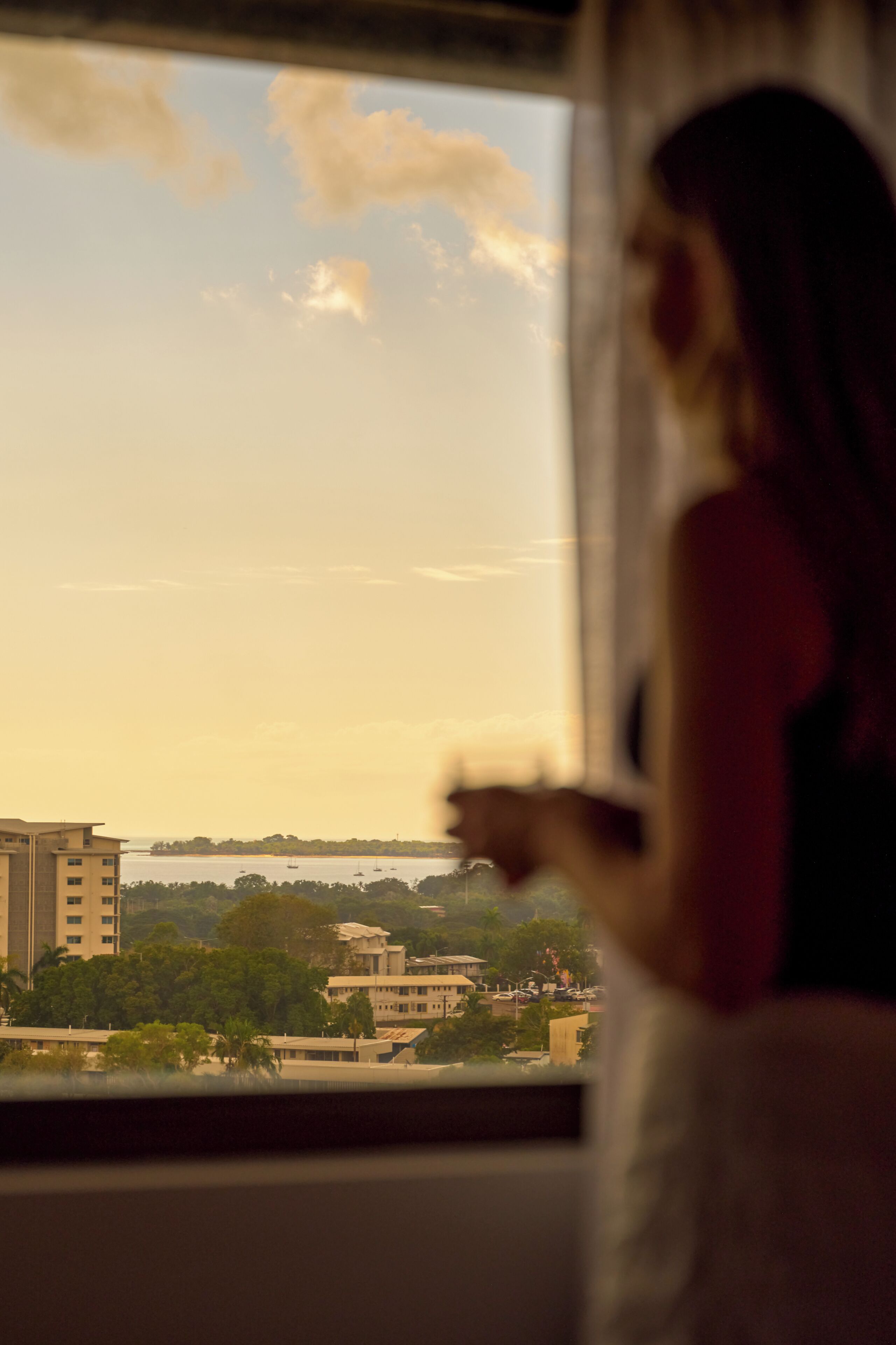 Eine Person steht an einem Fenster und blickt auf eine Stadtlandschaft mit Gebäuden und einem Gewässer.