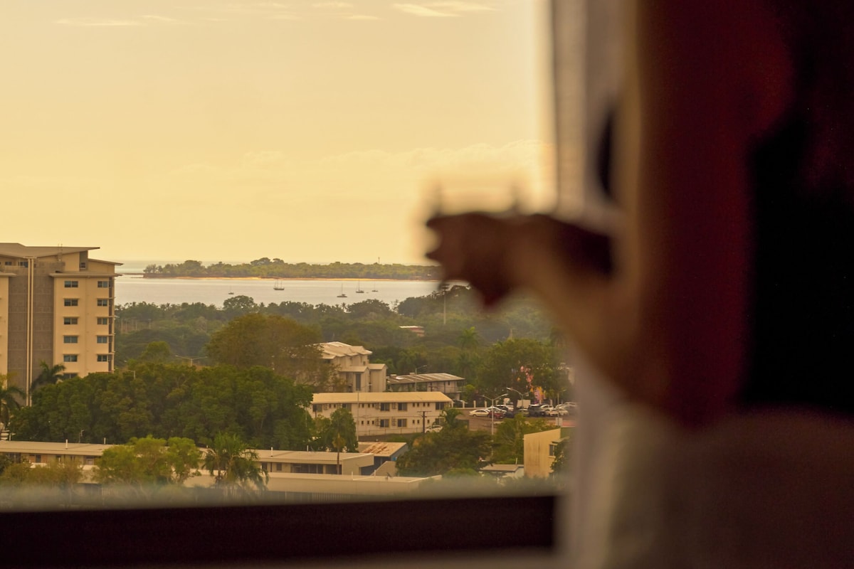 A person standing by a window overlooking a cityscape with buildings and a body of water.