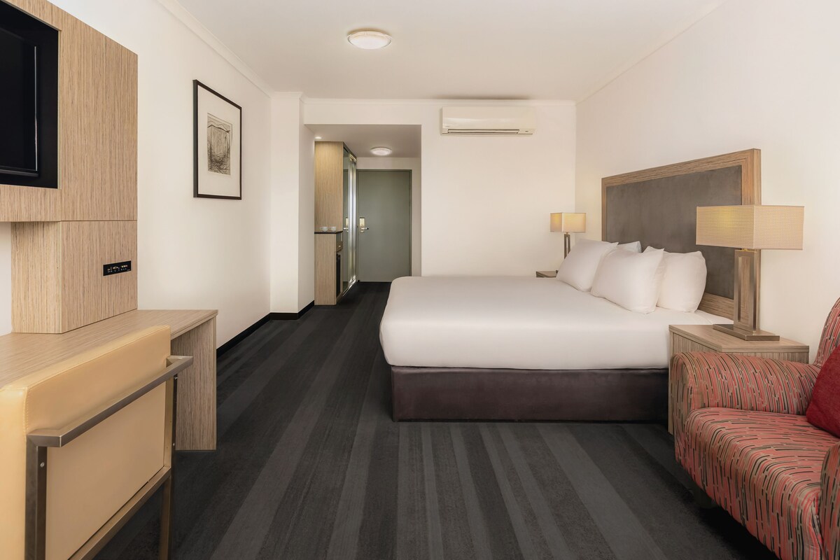 A hotel room with a bed, a desk, a chair, a TV, a door, and an air conditioning unit.