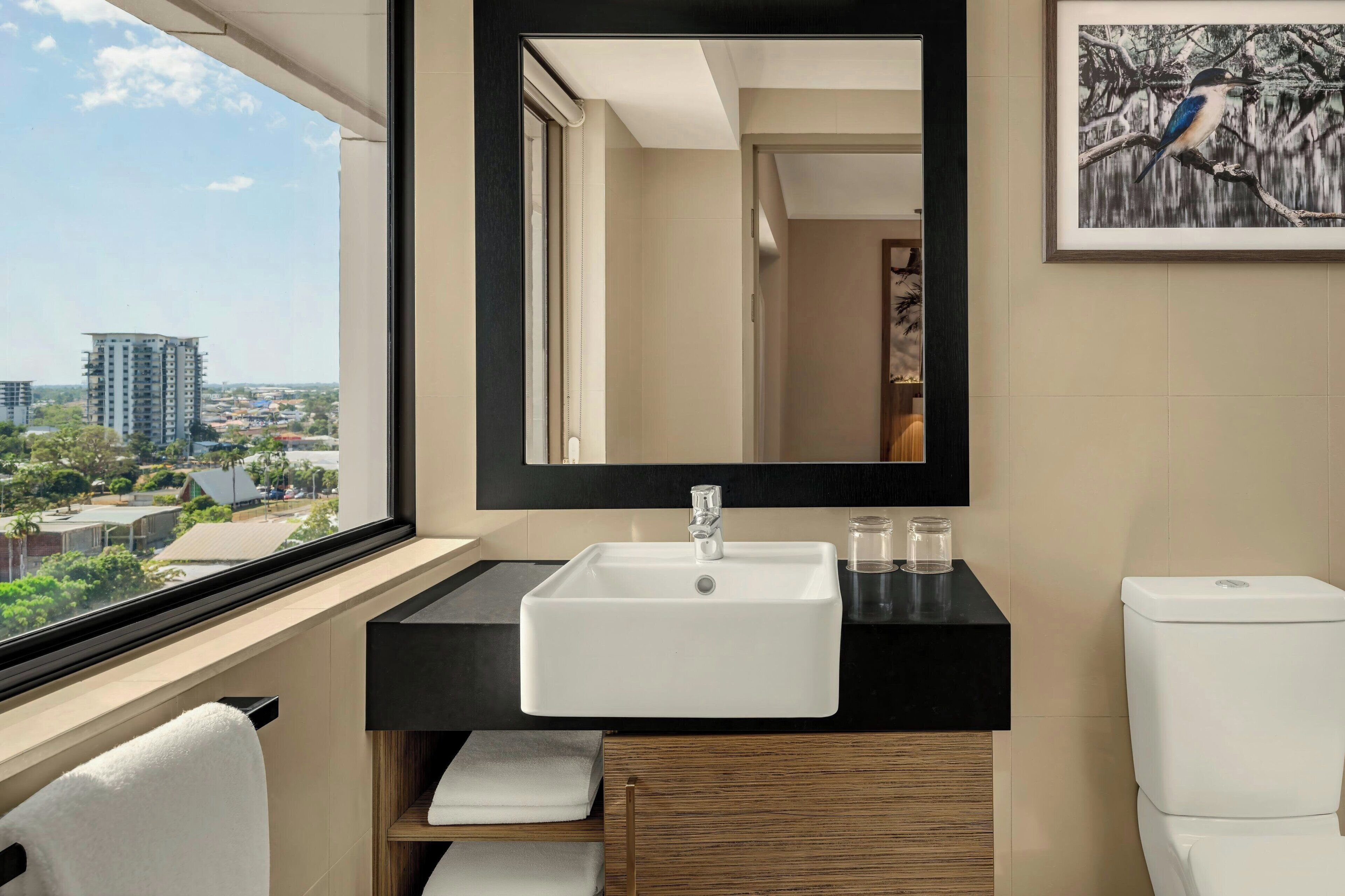 Un baño moderno con un ventanal grande, un lavabo blanco, una encimera negra, un armario de madera y un inodoro.